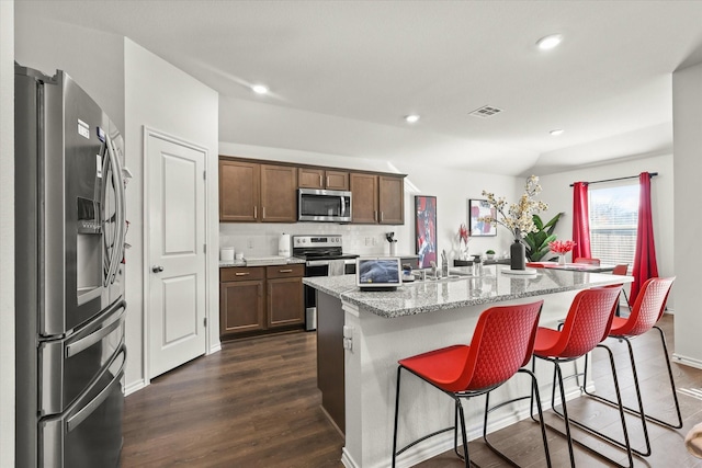 kitchen with dark wood-type flooring, stainless steel appliances, light stone counters, a center island with sink, and a breakfast bar area