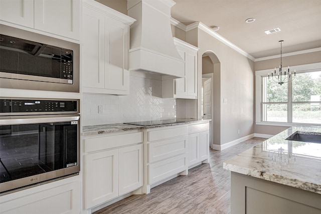 kitchen with white cabinetry, light stone countertops, stainless steel appliances, custom range hood, and a chandelier