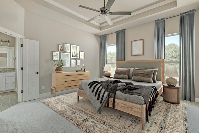 carpeted bedroom with ceiling fan, a raised ceiling, ensuite bathroom, and multiple windows