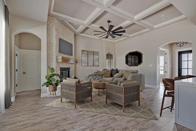 living room with ceiling fan, a towering ceiling, beamed ceiling, coffered ceiling, and a stone fireplace