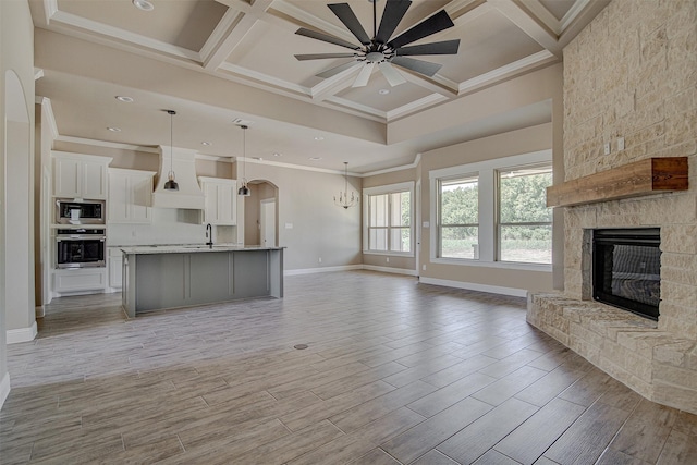 unfurnished living room with ceiling fan, sink, coffered ceiling, a stone fireplace, and ornamental molding