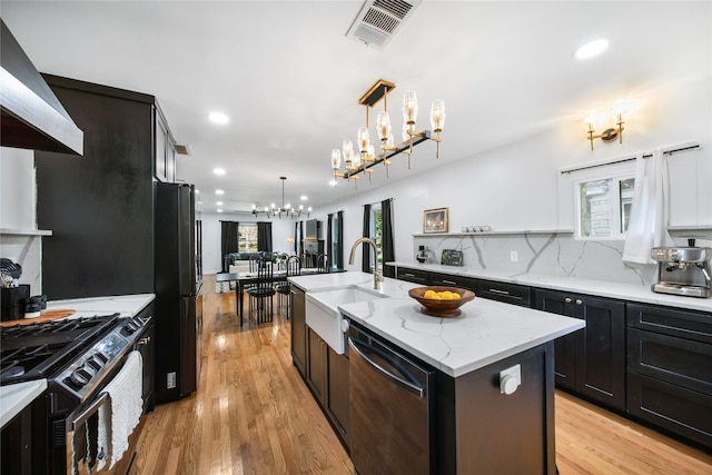 kitchen featuring black appliances, wall chimney exhaust hood, decorative light fixtures, sink, and a center island with sink