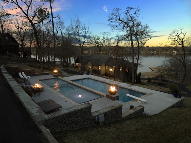 pool at dusk featuring a patio area, an in ground hot tub, an outdoor fire pit, a diving board, and a water view