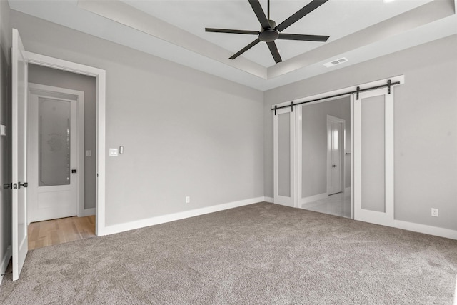 unfurnished bedroom with ceiling fan, a barn door, a tray ceiling, and light colored carpet