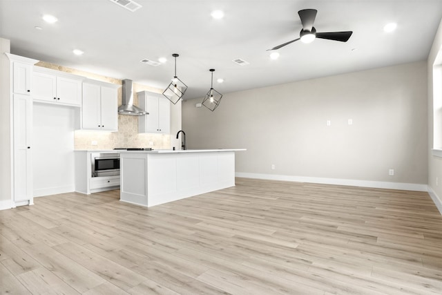 kitchen featuring wall chimney exhaust hood, a center island with sink, white cabinetry, and light countertops