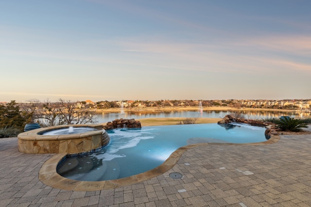 pool at dusk featuring a water view, pool water feature, a patio, and an in ground hot tub