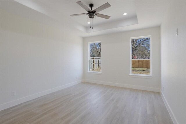 empty room with ceiling fan, a tray ceiling, light wood-type flooring, and a healthy amount of sunlight