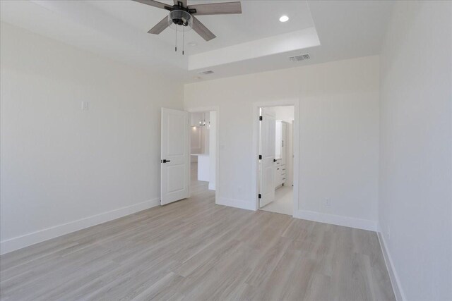 unfurnished bedroom with ceiling fan, light wood-type flooring, a tray ceiling, and connected bathroom