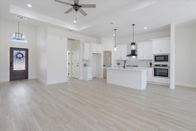 kitchen featuring white cabinetry, a center island with sink, stainless steel oven, built in microwave, and wall chimney exhaust hood