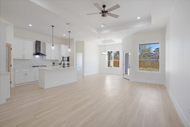 unfurnished living room with sink, light wood-type flooring, ceiling fan with notable chandelier, and a tray ceiling