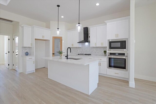 kitchen with wall chimney range hood, sink, a kitchen island with sink, appliances with stainless steel finishes, and white cabinets