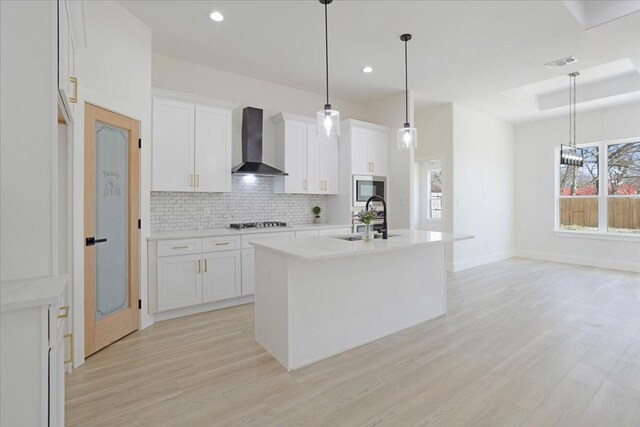 kitchen with an island with sink, hanging light fixtures, wall chimney exhaust hood, white cabinets, and sink