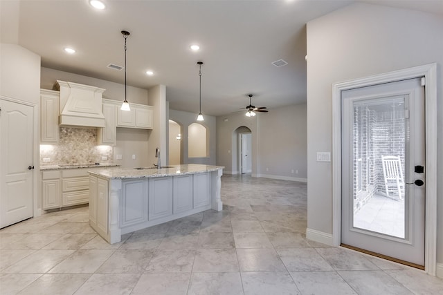 kitchen with premium range hood, an island with sink, ceiling fan, decorative light fixtures, and light stone countertops