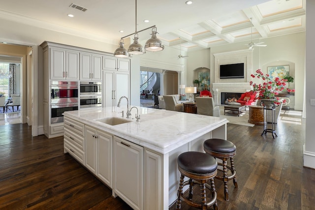 kitchen with decorative light fixtures, stainless steel double oven, a center island with sink, and coffered ceiling