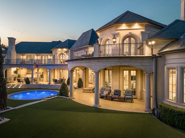 back house at dusk featuring a balcony, a patio area, and a lawn