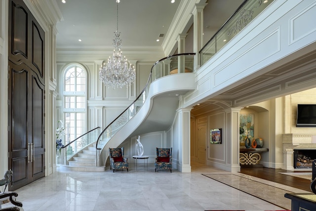 foyer entrance featuring a towering ceiling, a premium fireplace, an inviting chandelier, crown molding, and decorative columns