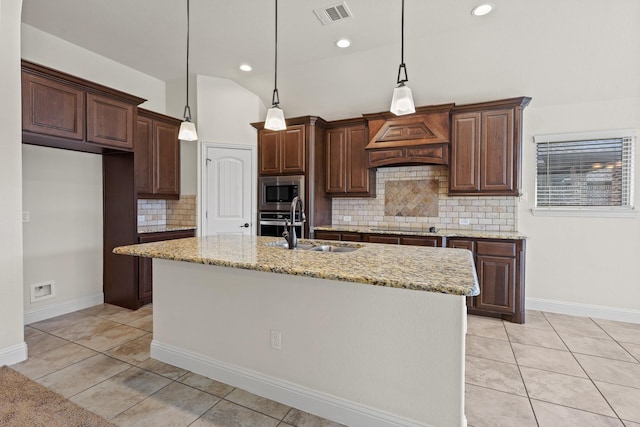 kitchen with light stone countertops, pendant lighting, stainless steel appliances, premium range hood, and a kitchen island with sink