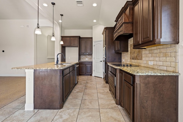 kitchen with light stone countertops, dark brown cabinetry, sink, hanging light fixtures, and a kitchen island with sink