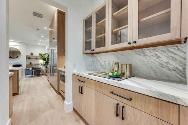 bar featuring backsplash, high end refrigerator, light brown cabinetry, light stone countertops, and light hardwood / wood-style flooring