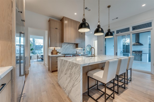 kitchen with hanging light fixtures, a kitchen island with sink, light wood-type flooring, wall chimney exhaust hood, and light stone counters