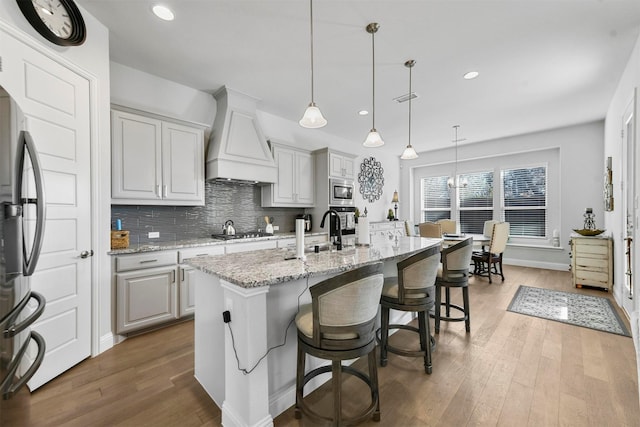 kitchen with a center island with sink, stainless steel appliances, hanging light fixtures, custom range hood, and light stone counters