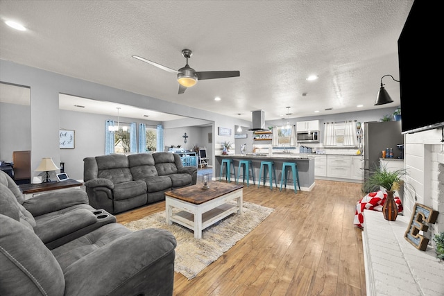 living room featuring ceiling fan with notable chandelier, light hardwood / wood-style floors, a textured ceiling, and a brick fireplace