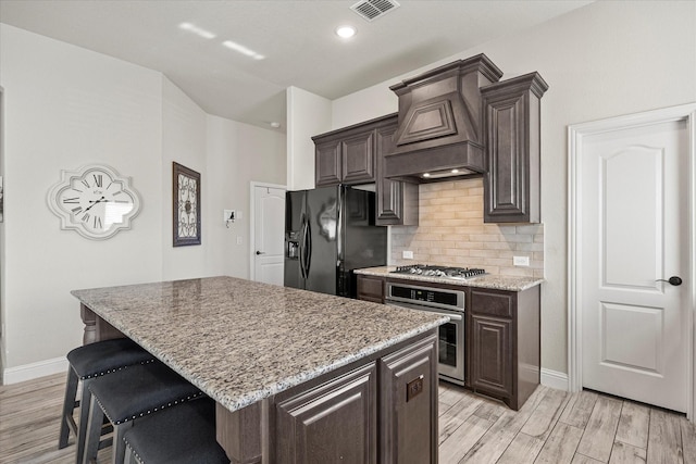 kitchen featuring dark brown cabinetry, appliances with stainless steel finishes, custom exhaust hood, a kitchen island, and light hardwood / wood-style flooring