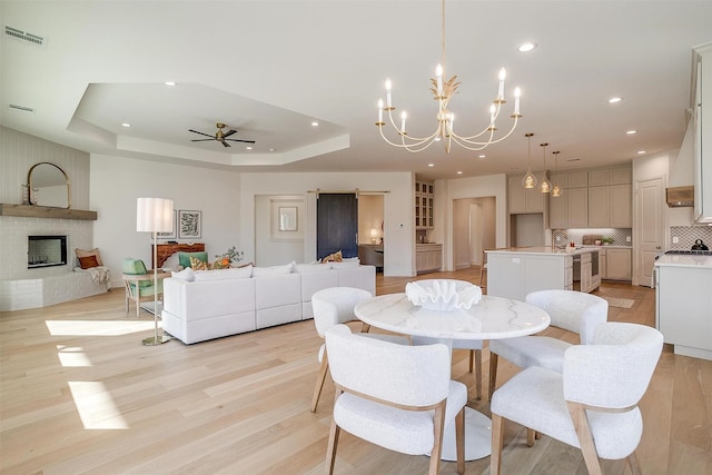 dining space featuring a raised ceiling, a fireplace, ceiling fan with notable chandelier, and light hardwood / wood-style flooring