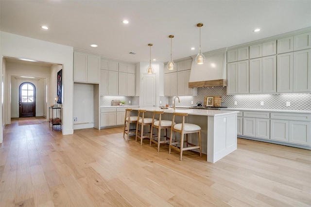 kitchen with premium range hood, light hardwood / wood-style flooring, hanging light fixtures, a kitchen island with sink, and a breakfast bar area