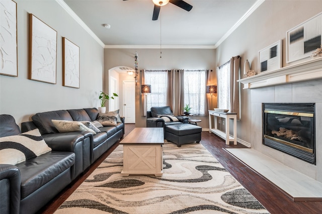 living area with arched walkways, a ceiling fan, a tile fireplace, dark wood-style flooring, and crown molding