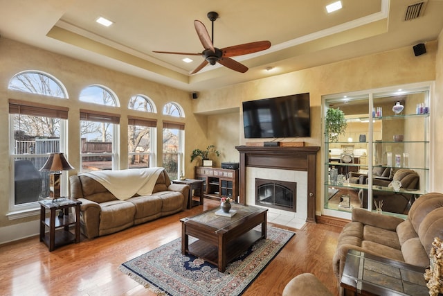 living room featuring ceiling fan, a tiled fireplace, light hardwood / wood-style flooring, and a raised ceiling