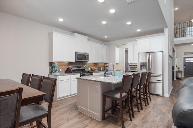 kitchen featuring white cabinetry, appliances with stainless steel finishes, a kitchen island with sink, light stone countertops, and light hardwood / wood-style flooring