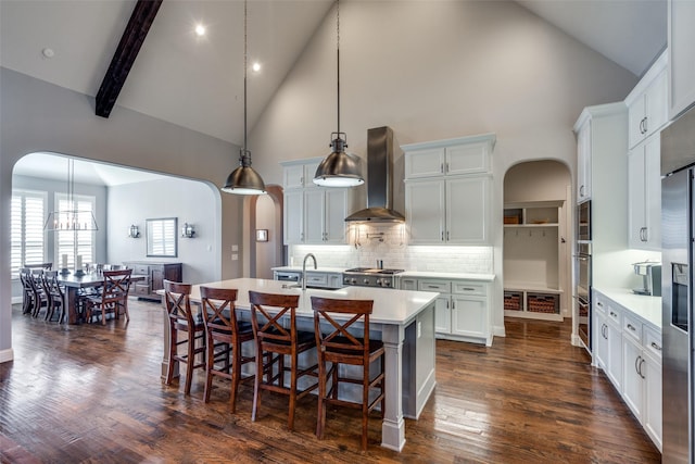 kitchen with backsplash, a kitchen island with sink, hanging light fixtures, high vaulted ceiling, and wall chimney exhaust hood