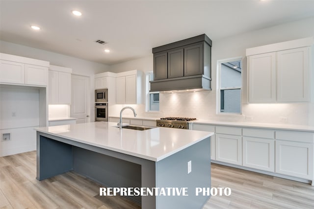 kitchen with white cabinets, stainless steel appliances, light hardwood / wood-style floors, sink, and a center island with sink