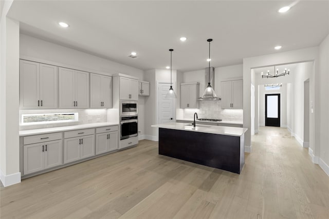 kitchen featuring hanging light fixtures, a kitchen island with sink, stainless steel appliances, light countertops, and wall chimney range hood