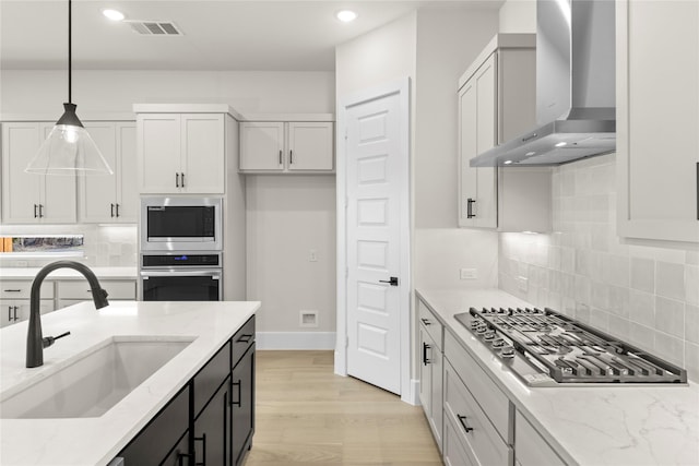 kitchen featuring pendant lighting, stainless steel appliances, a sink, light stone countertops, and wall chimney exhaust hood