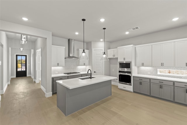 kitchen featuring sink, white cabinetry, an island with sink, stainless steel appliances, and wall chimney range hood