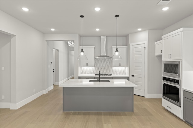 kitchen with sink, stainless steel oven, an island with sink, decorative light fixtures, and wall chimney exhaust hood