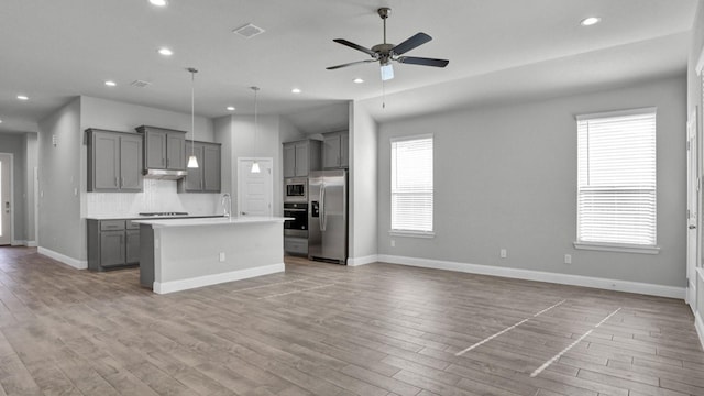 kitchen with light hardwood / wood-style floors, ceiling fan, stainless steel appliances, backsplash, and a kitchen island with sink