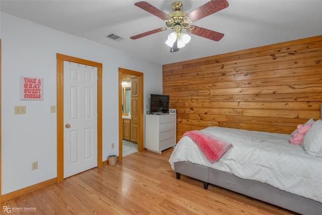 bedroom featuring ceiling fan, light hardwood / wood-style floors, ensuite bathroom, and wooden walls