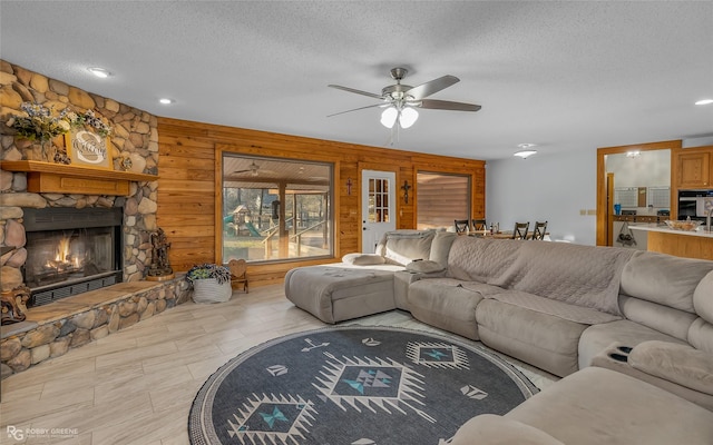 living room featuring ceiling fan, a stone fireplace, wooden walls, and a textured ceiling