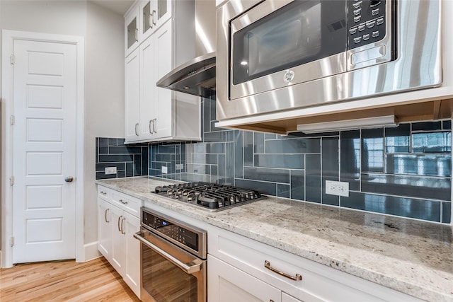 kitchen with white cabinets, wall chimney range hood, stainless steel appliances, backsplash, and light hardwood / wood-style flooring