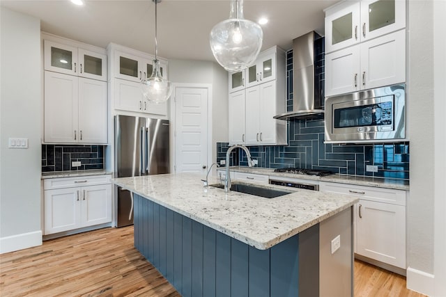 kitchen with stainless steel appliances, a kitchen island with sink, wall chimney range hood, pendant lighting, and white cabinets