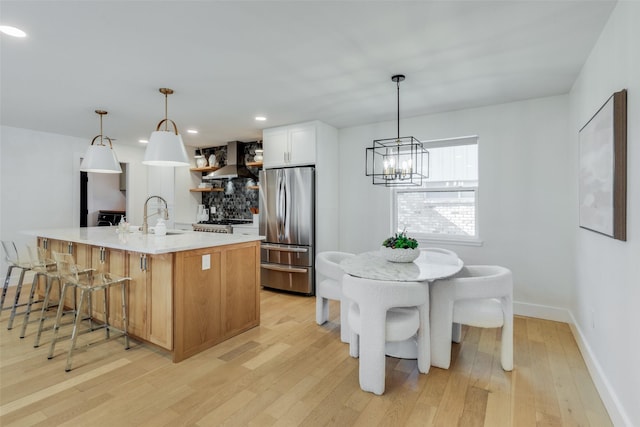 kitchen featuring a large island with sink, wall chimney exhaust hood, decorative light fixtures, white cabinetry, and stainless steel fridge