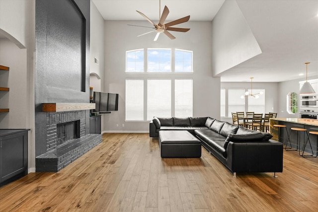 living room with light wood-type flooring, a brick fireplace, ceiling fan with notable chandelier, and a high ceiling