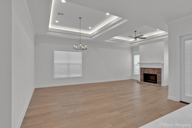 unfurnished living room with light hardwood / wood-style floors, a brick fireplace, a raised ceiling, ornamental molding, and ceiling fan with notable chandelier