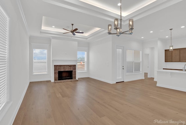 unfurnished living room featuring a raised ceiling, a brick fireplace, a wealth of natural light, and ceiling fan with notable chandelier