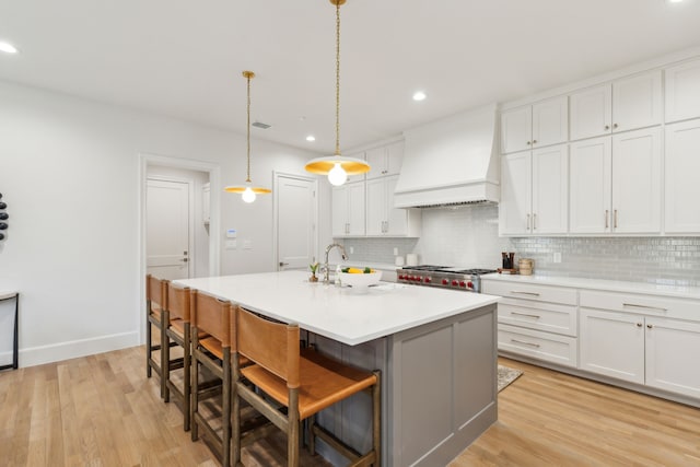 kitchen featuring premium range hood, white cabinetry, a center island with sink, a breakfast bar area, and decorative light fixtures