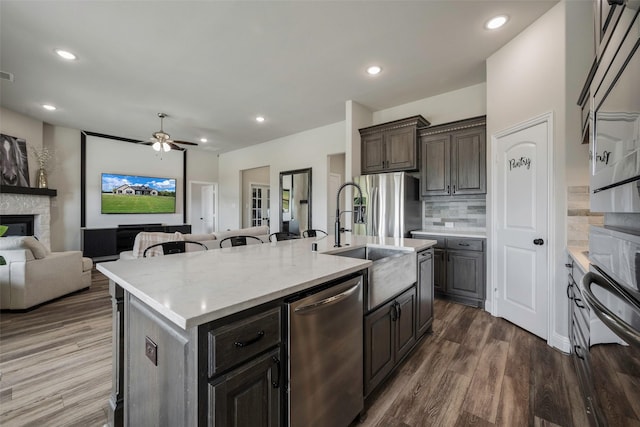 kitchen featuring appliances with stainless steel finishes, a fireplace, backsplash, ceiling fan, and a center island with sink