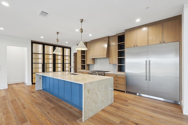 kitchen with custom exhaust hood, a center island with sink, appliances with stainless steel finishes, hanging light fixtures, and light stone counters